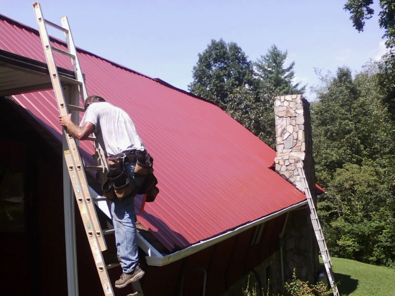 Roofer on a ladder beside a red metal roof and stone chimney — Soffit & Fascia Repair by Roofing Brothers in Saxapahaw
