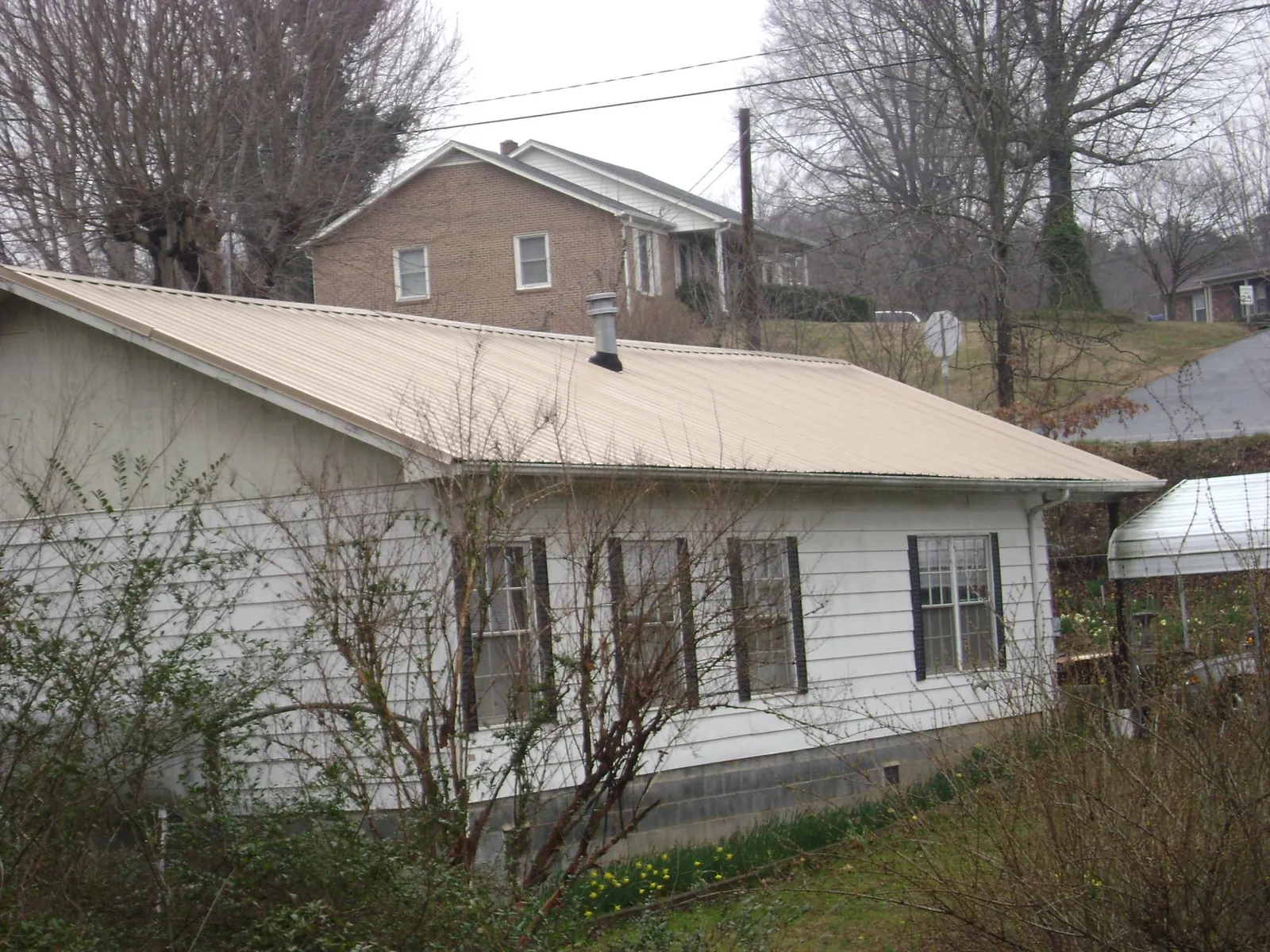 Tan metal roof on a white sided house set into a hillside — Commercial Roofing by Roofing Brothers in Saxapahaw